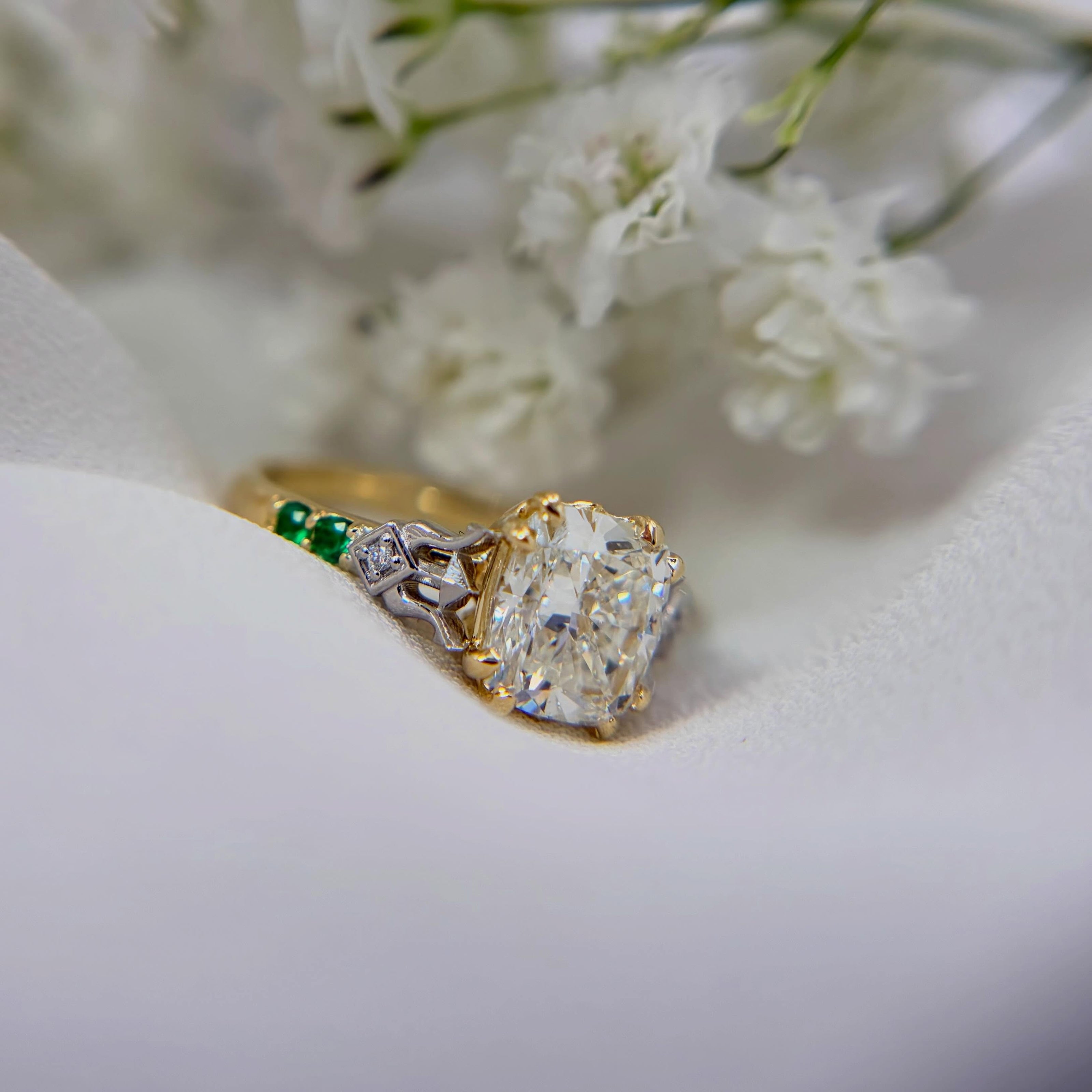 A yellow gold ring with a large cushion-cut clear center stone and green emerald accent stones on the band resting on soft white fabric with blurred white flowers in the background