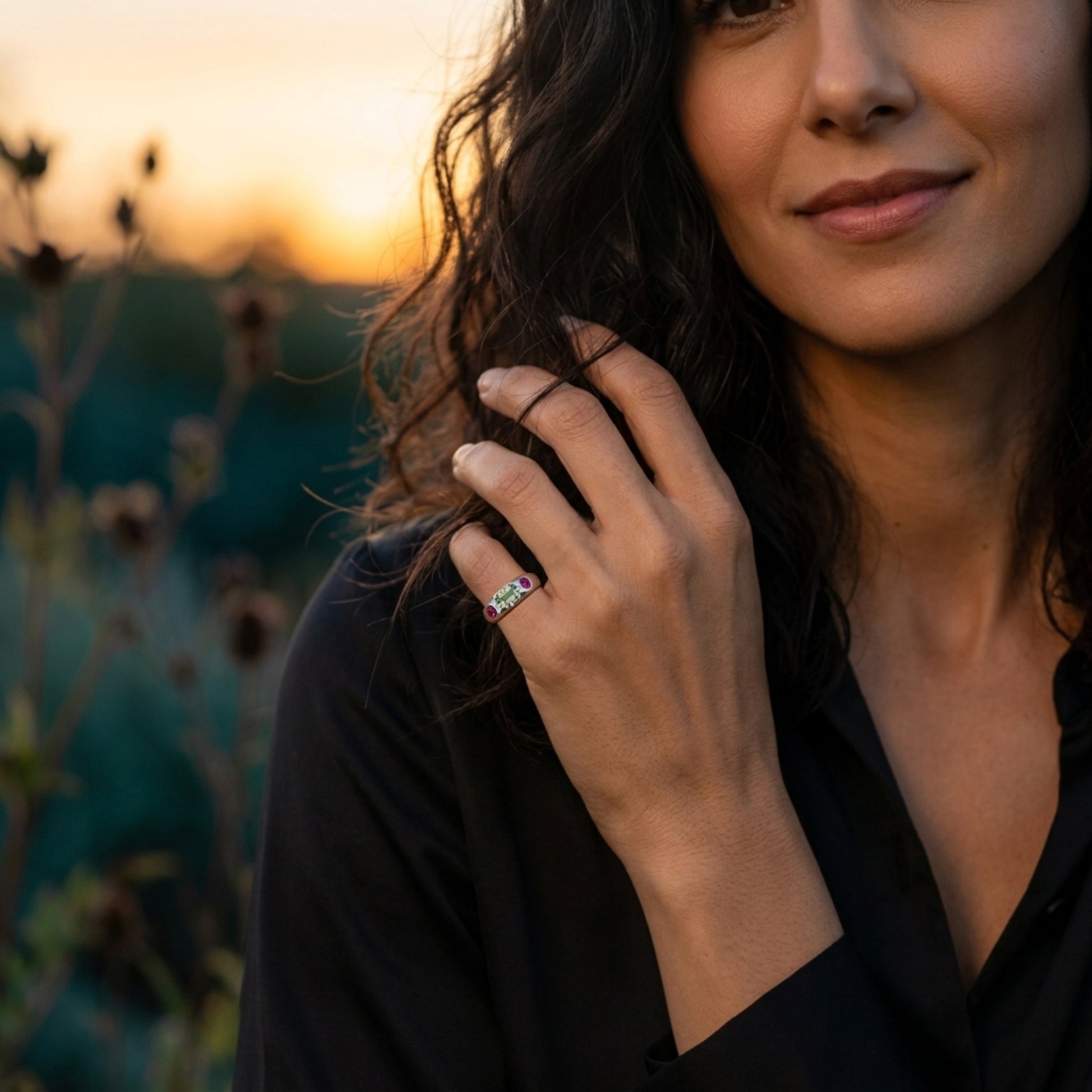 A close-up lifestyle shot of a woman wearing a white band ring with small colorful gemstones on her finger, set against a warm sunset background.