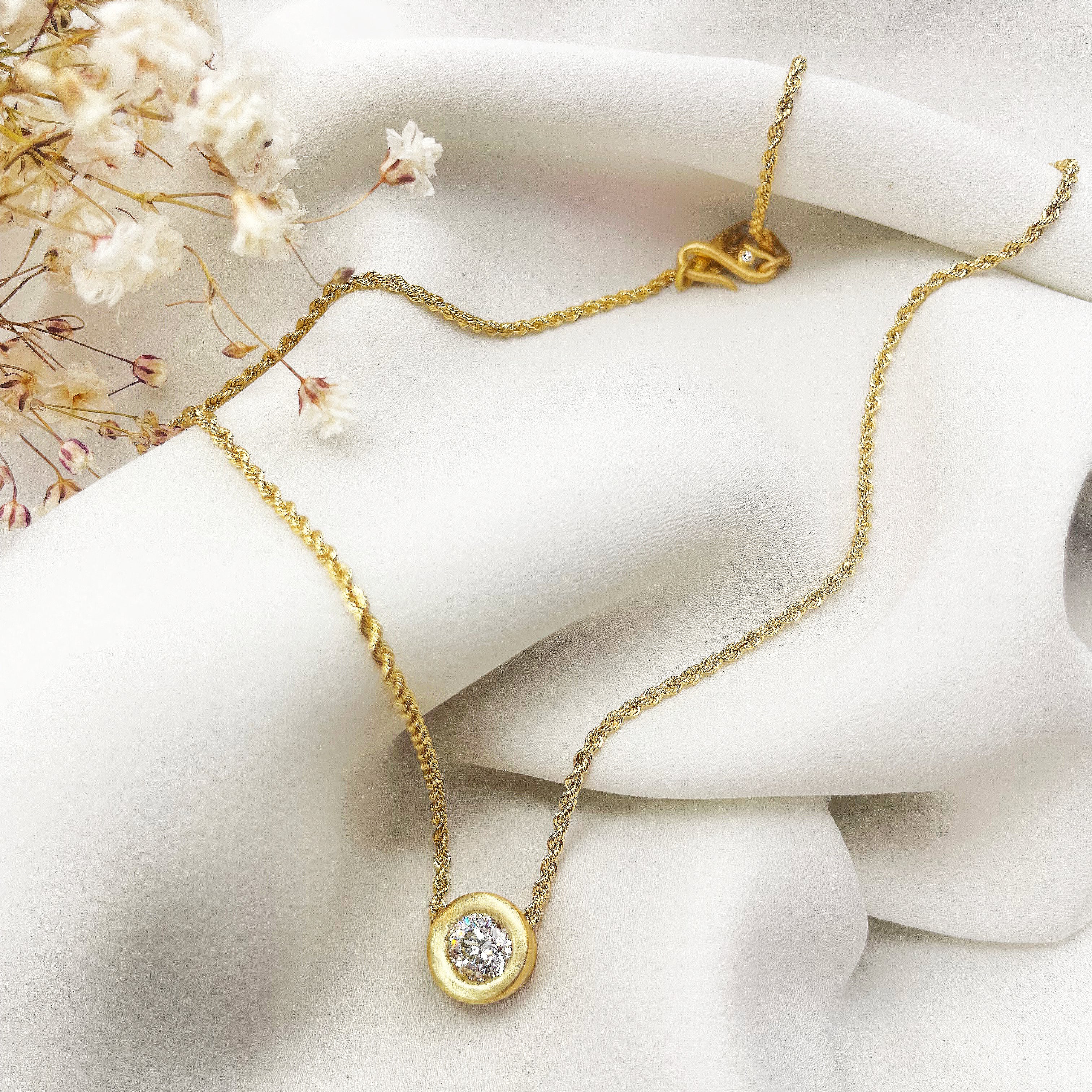 Gold necklace with a diamond pendant on a white surface with dried flowers.