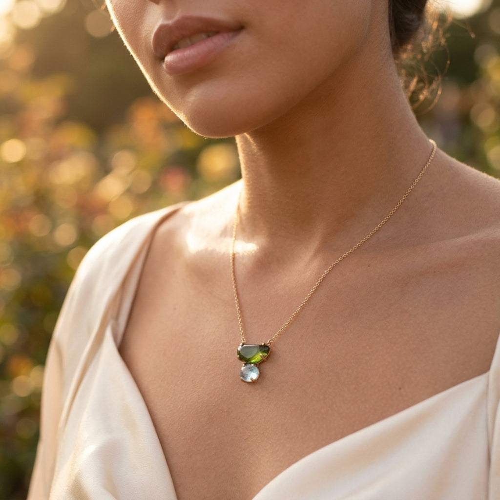 Woman wearing a gold necklace with a green tourmaline and blue topaz gemstone pendant against a blurred natural background