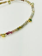 A close-up of a beaded strand jewelry piece featuring faceted beads in green, pink, yellow, and gray, accented with small gold-toned beads, on a plain light background.