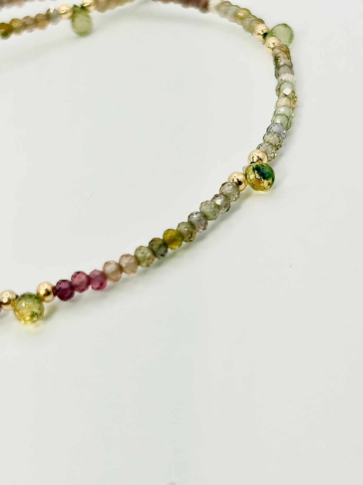 A close-up of a beaded strand jewelry piece featuring faceted beads in green, pink, yellow, and gray, accented with small gold-toned beads, on a plain light background.