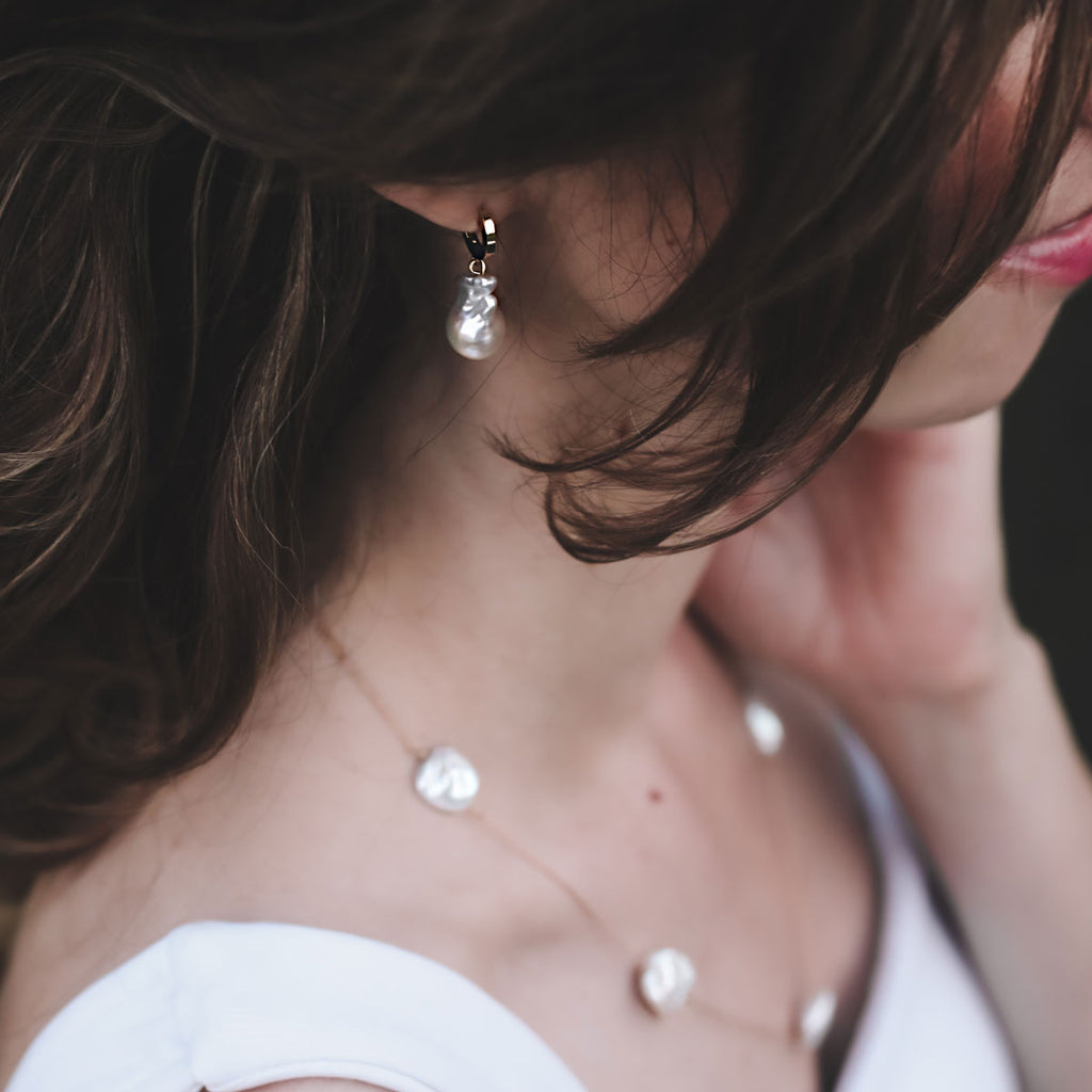 Close-up of a person wearing baroque pearl huggie hoop earrings and a necklace with a blurred background