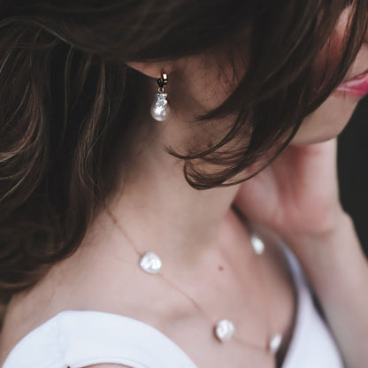 Close-up of a person wearing baroque pearl huggie hoop earrings and a necklace with a blurred background