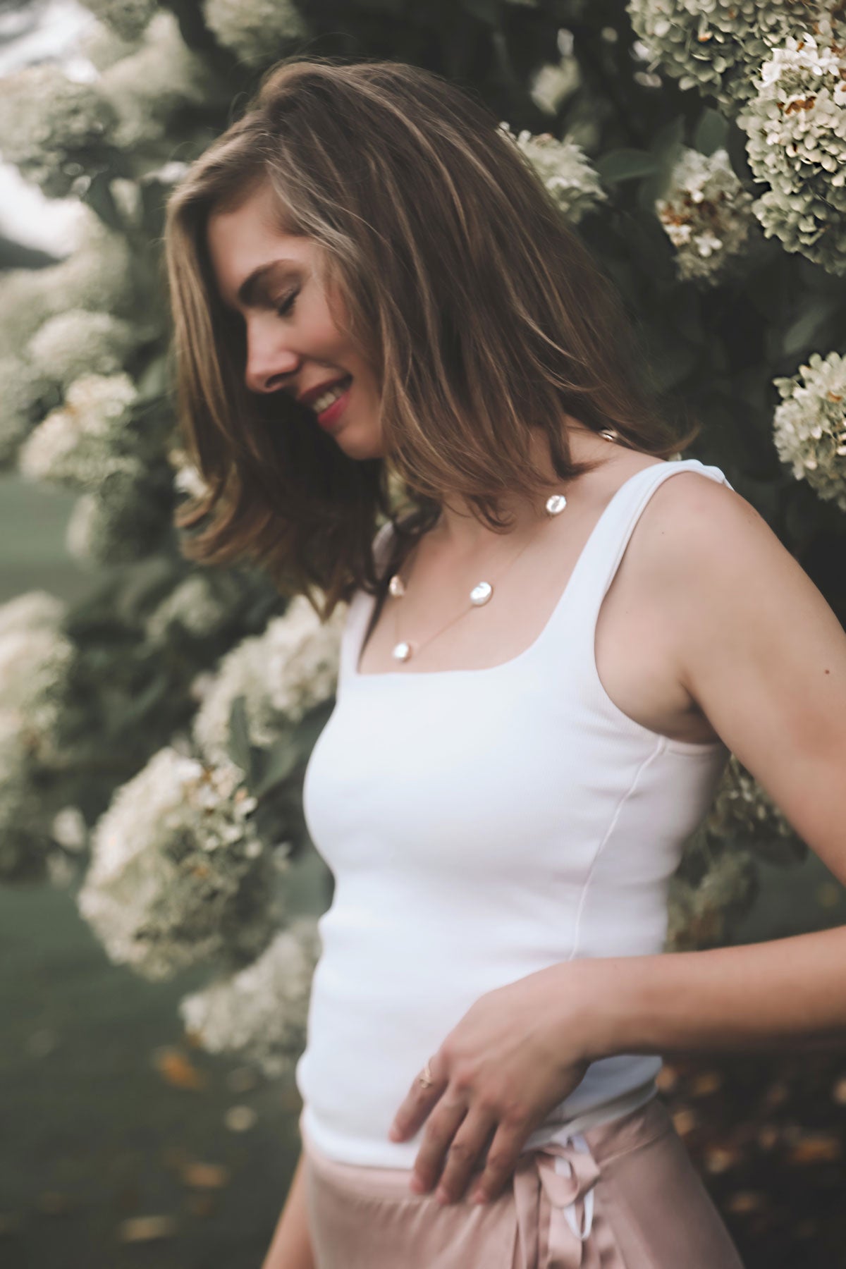 Woman wearing a white tank top and haven keshi pearl station necklace in 14K yellow gold with a blurred floral background