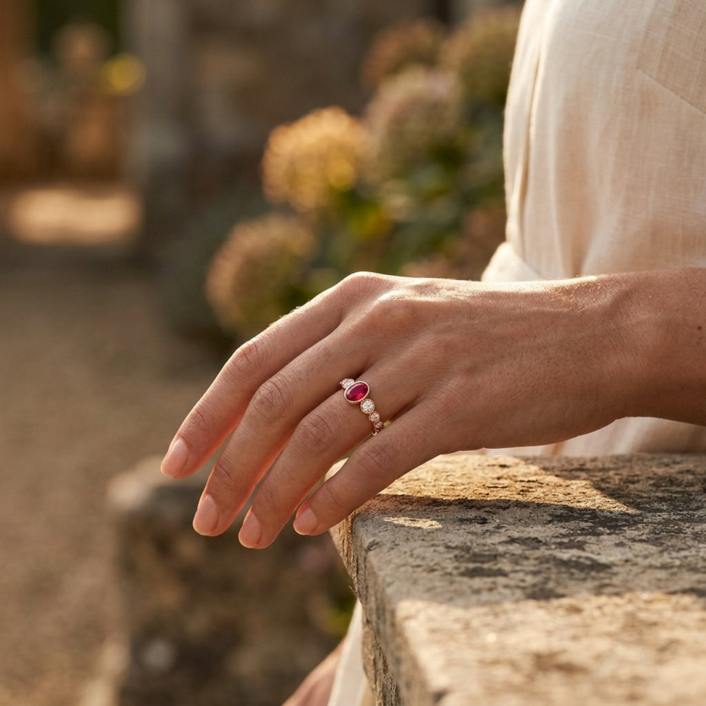 Hand wearing a ring with a pinkish red ruby gemstone accented by rose gold bezel set diamonds  on a stone surface with a blurred natural background
