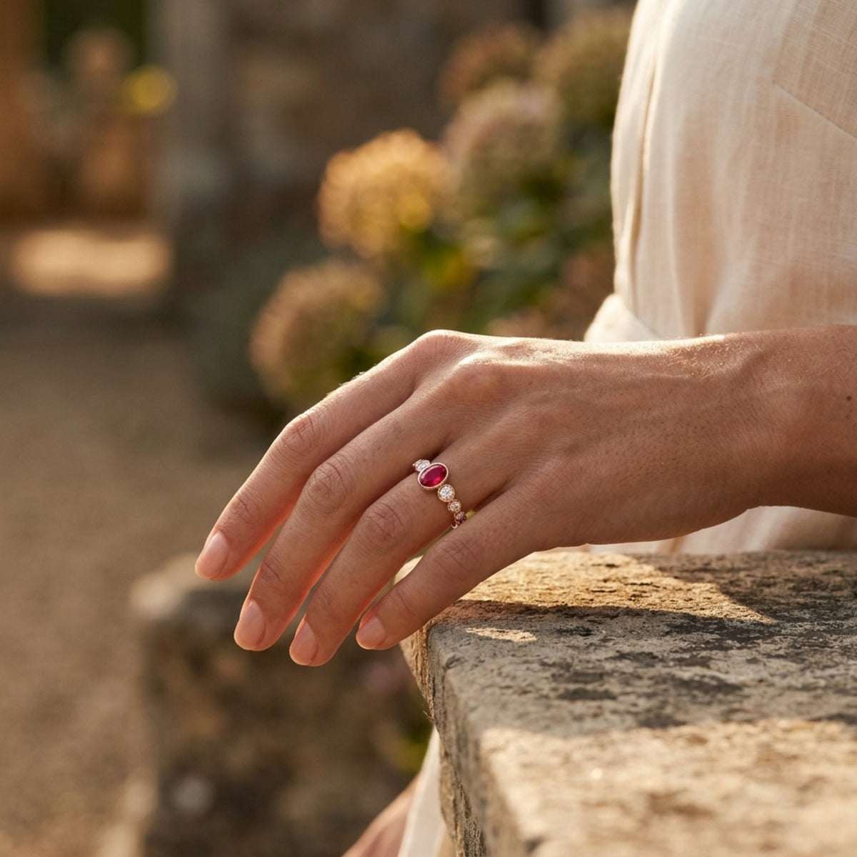 Hand wearing a ring with a pinkish red ruby gemstone accented by rose gold bezel set diamonds  on a stone surface with a blurred natural background