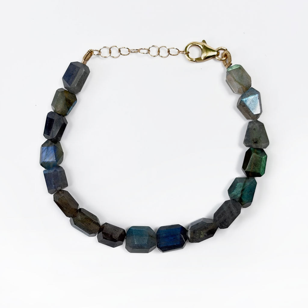 A beaded bracelet made of faceted dark blue and gray labradorite stones, connected by a gold-tone chain extender and lobster clasp, shown on a plain white background.