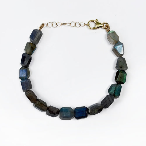 A beaded bracelet made of faceted dark blue and gray labradorite stones, connected by a gold-tone chain extender and lobster clasp, shown on a plain white background.