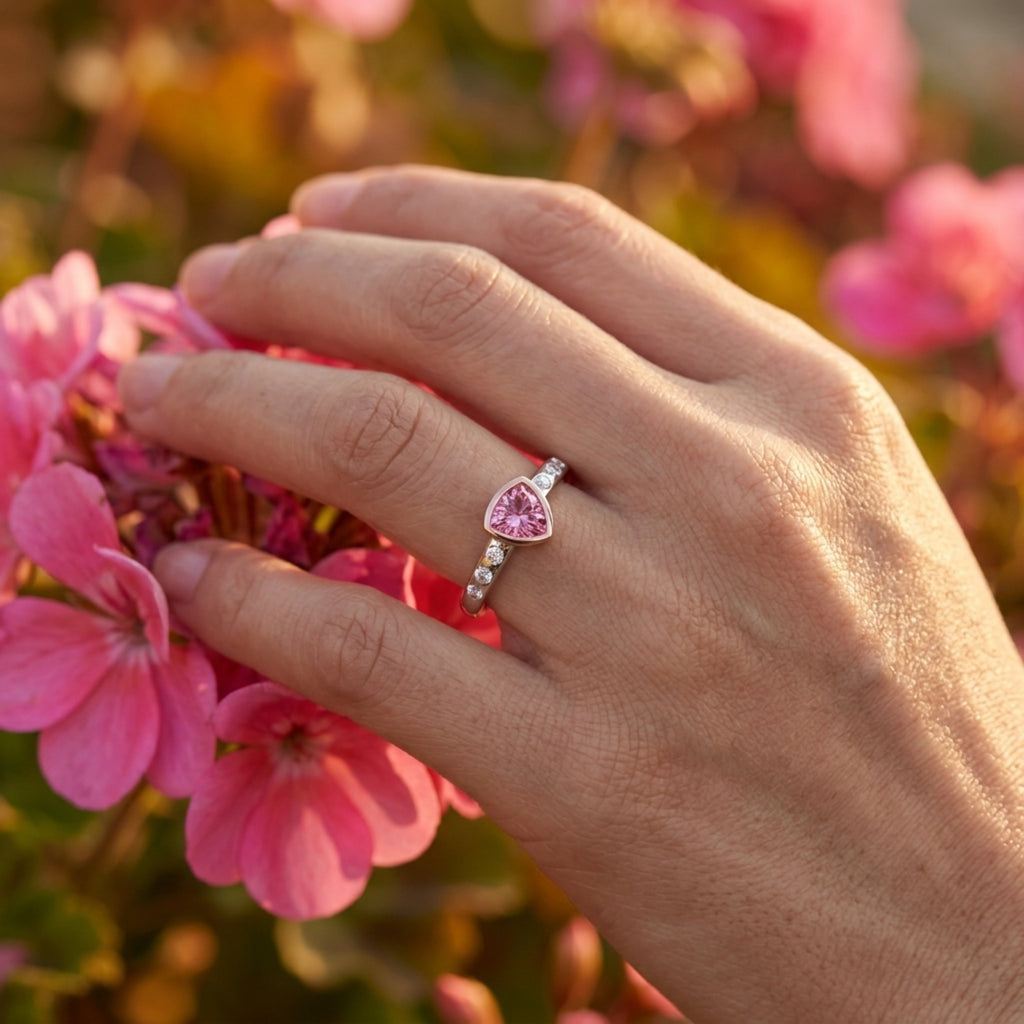 Hand wearing a pink trillion-shaped gemstone ring bezel set in rose gold with white gold shank and accent diamonds with pink flowers in the background