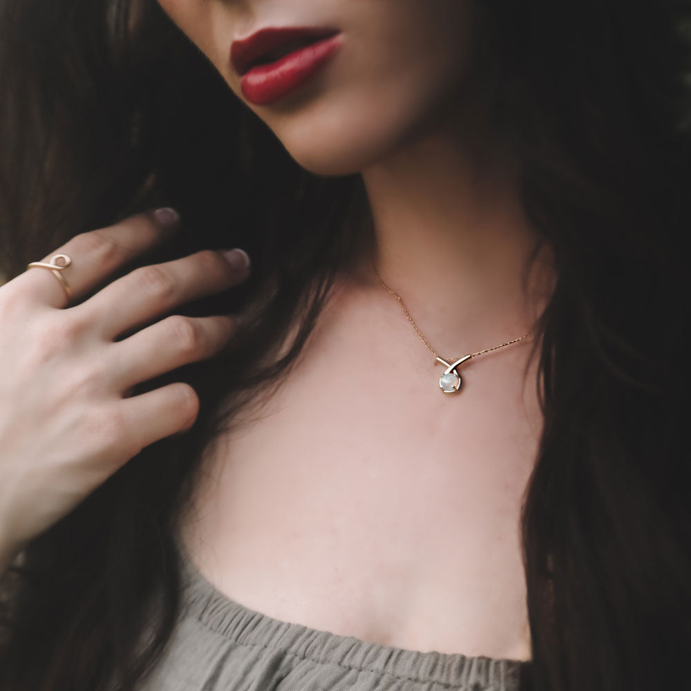 Close-up of a woman wearing a gold necklace with a small loop pendant featuring a natural ethiopian opal gemstone, hand touching hair, on a dark background