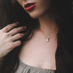 Close-up of a woman wearing a gold necklace with a small loop pendant featuring a natural ethiopian opal gemstone, hand touching hair, on a dark background