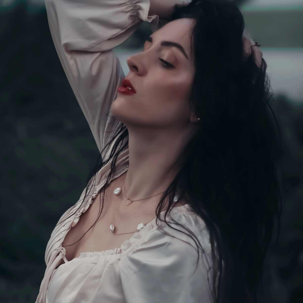 haven station necklace and fish scales necklace worn by model with red lip, black wispy hair and silk off white blouse near dusk in an outdoor setting