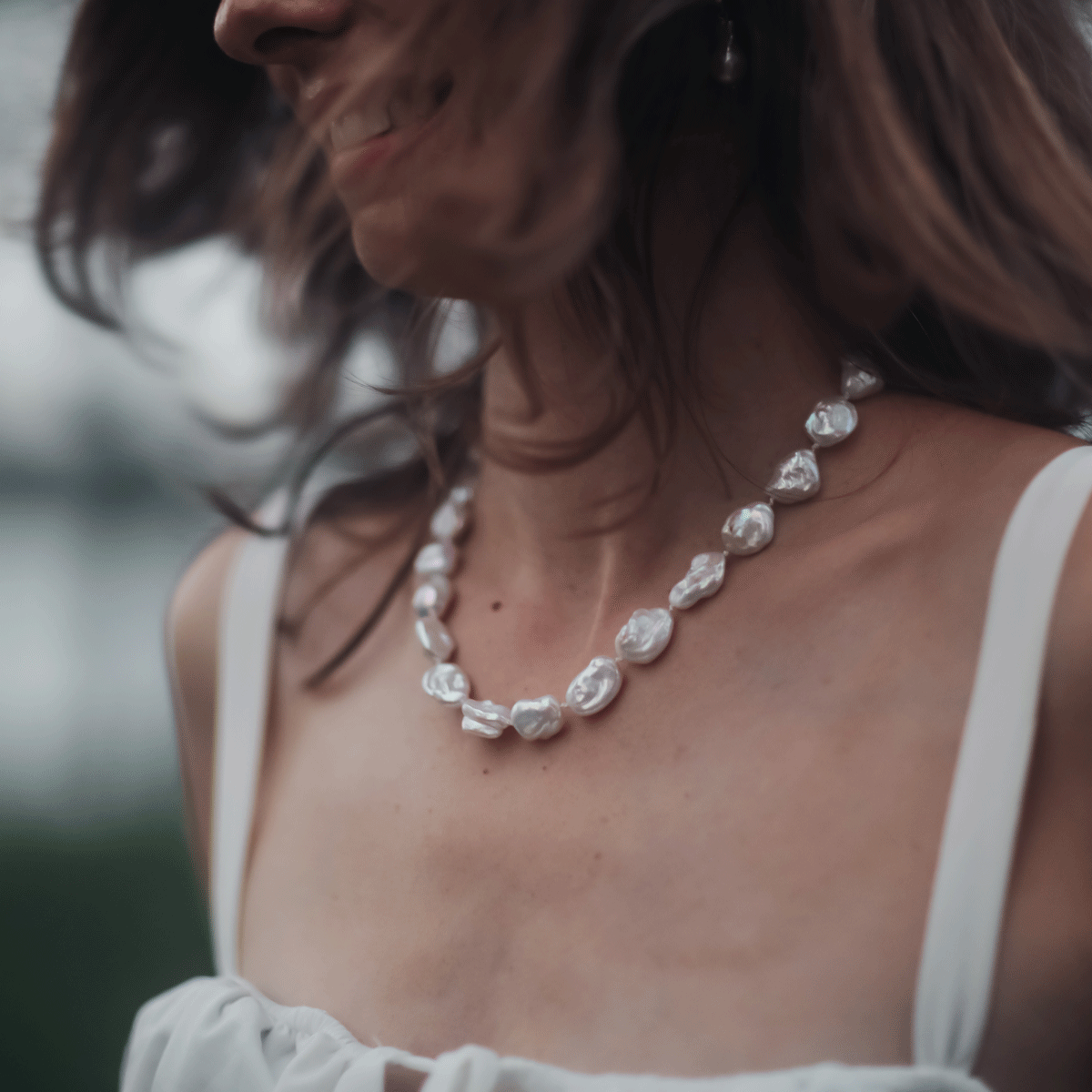 keshi pearl strand necklace worn by woman in white dress with hair blowing in the wind against a natural background