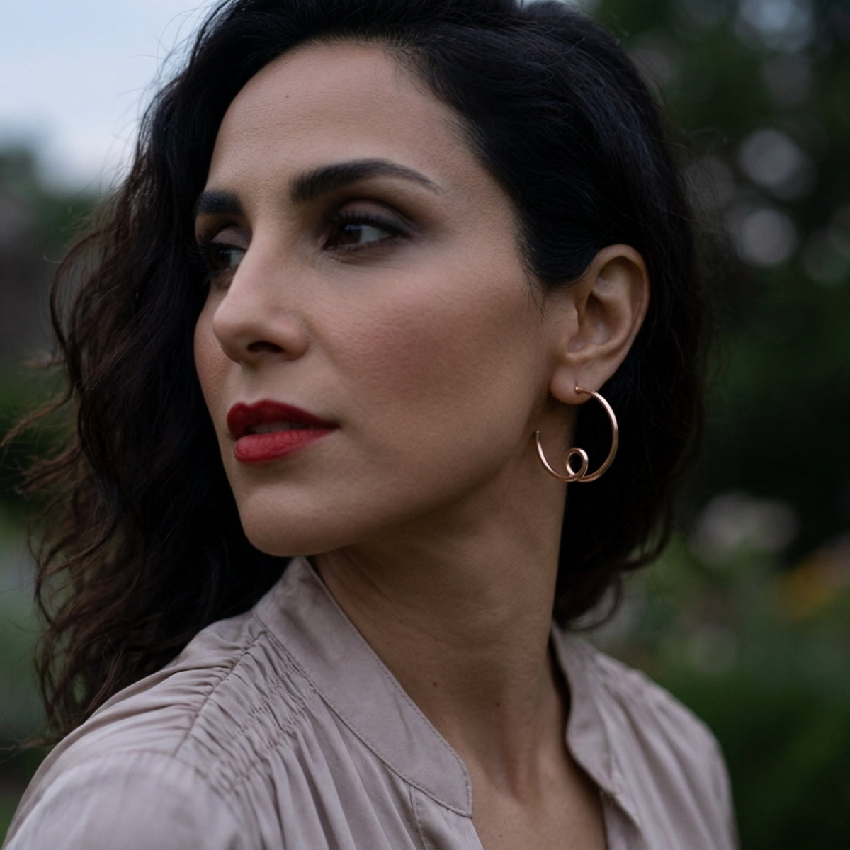 Woman with dark hair and red lipstick wearing rose gold large flow hoop earrings by Jillery, outdoors.