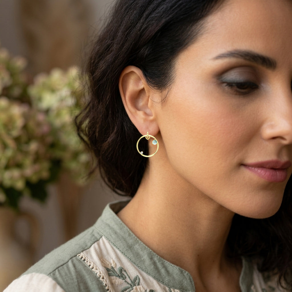 Close-up of a woman wearing yellow gold hoop earrings accented by blue topaz cabochon gemstones and a white diamond with a blurred floral background