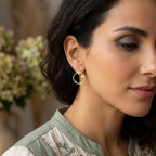 Close-up of a woman wearing yellow gold hoop earrings accented by blue topaz cabochon gemstones and a white diamond with a blurred floral background