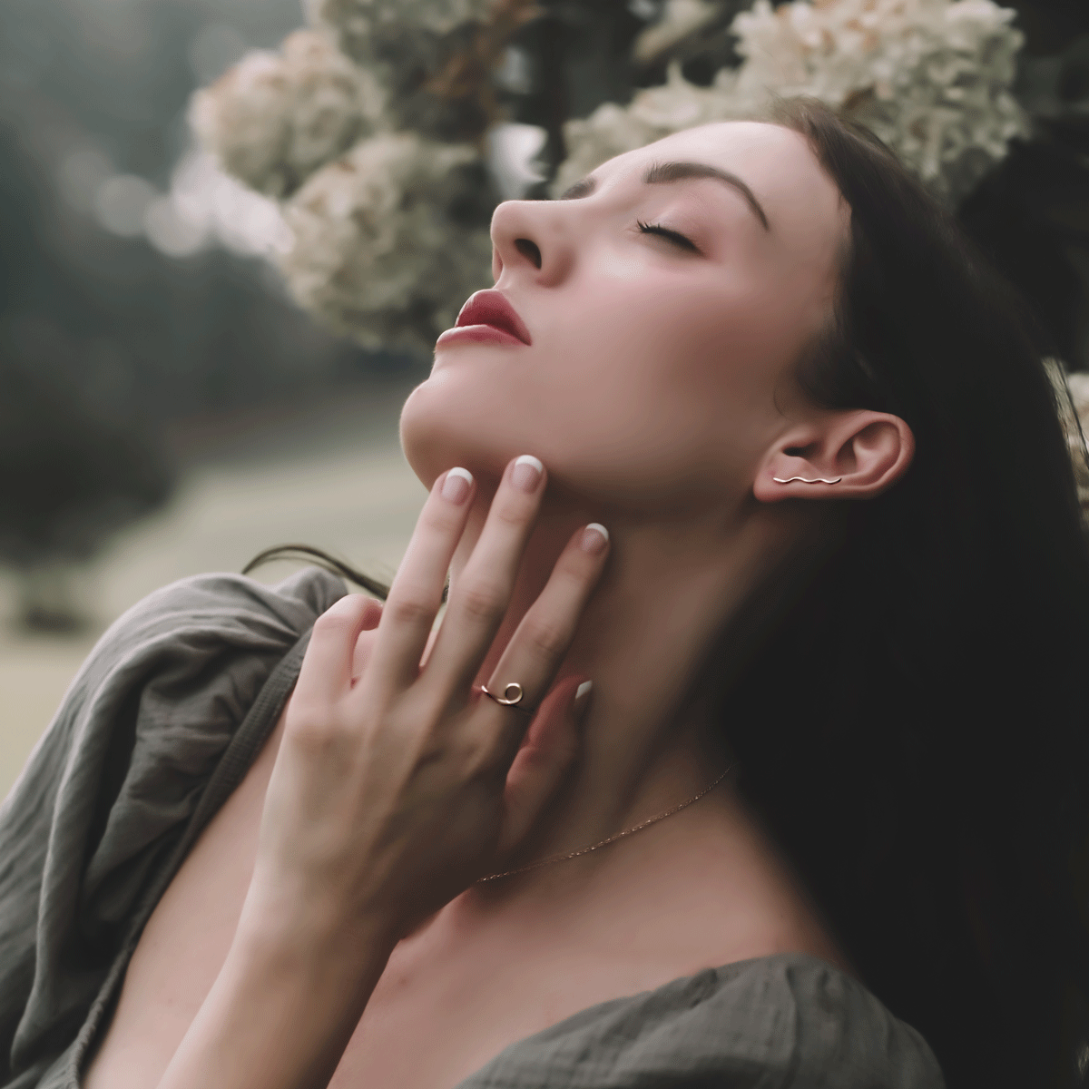 beautiful and dramatic pose of woman tilting head back with eyes closed featuring 14K yellow gold squiggle ear climber and yellow gold flow ring in an outdoor setting with white flowers blurred in the background